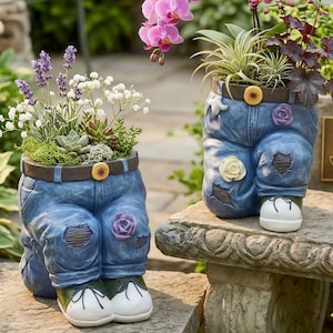 Puede incluir: Dos macetas decorativas con forma de piernas de mezclilla, cada una con un cinturón marrón y un botón amarillo. Las macetas están llenas de plantas y flores. Se encuentran sobre una superficie de piedra, creando un ambiente de jardín.