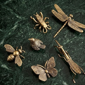 Könnte beinhalten: Eine Sammlung antiker Insekten- und Vogelfiguren aus Messing. Das Set umfasst eine Libelle, eine Biene, einen Schmetterling, eine Heuschrecke, einen Vogel und eine Grille. Die Figuren sind auf einer dunkelgrünen Marmoroberfläche angeordnet.