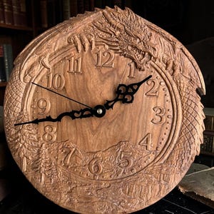 May include: A wooden wall clock with a dragon design carved around the face. The clock features black hands and Roman numerals. The background includes a mountain and forest scene.