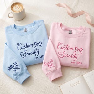 Może przedstawiać: Jasnoniebieska bluza i jasnoróżowa bluza, każda z wyhaftowanym napisem Custom Sorority est. 1895 oraz ikoną kokardki. Na rękawach wyhaftowano imiona Olivia i Sarah, każde z nich ozdobione jest dodatkowo małą kokardką.