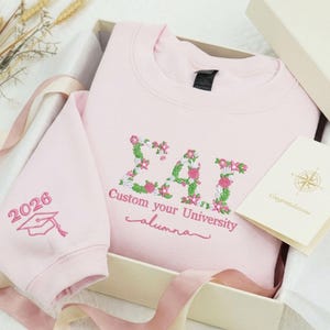 May include: A light pink sweatshirt with floral embroidery spelling out "ΣΑΙ" and the words "Custom your University alumna". The left sleeve has "2026" and a graduation cap. The sweatshirt is in a gift box with a card.