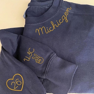 Op de afbeelding: Marineblauw sweatshirt met goud geborduurde tekst en ontwerpen. Het woord "Michigan" is op de borst geborduurd. De linkermouw heeft "USC" en een vredesteken. Een hart met het nummer "26" staat aan de linkerkant.