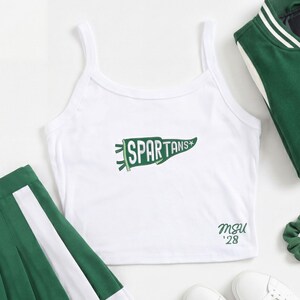 Pode incluir: Um top curto branco sem mangas com um gráfico de flâmula verde contendo o texto SPARTANS e uma pequena estrela. No canto inferior direito, exibe o texto verde MSU '28. A peça possui alças finas e um decote redondo simples.