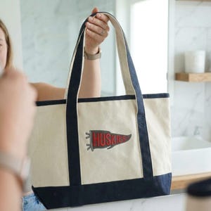 May include: Cream-colored tote bag with navy blue trim and handles. A red and gray pennant with the word "Huskies" is embroidered on the front. The bag is held up by a person.