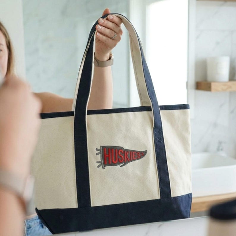 May include: Cream-colored tote bag with navy blue trim and handles. A red and gray pennant with the word "Huskies" is embroidered on the front. The bag is held up by a person.