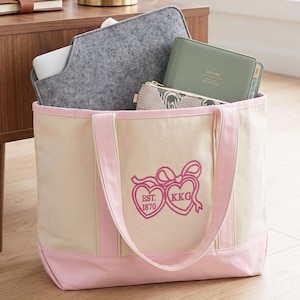 Puede incluir: Un bolso de lona color crema con ribetes y asas rosas, con un bordado rosa de dos corazones unidos por un lazo, con el texto EST. 1870 y KKG. El bolso contiene una funda de fieltro gris para portátil y una agenda de color verde.