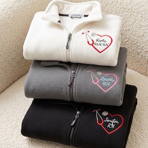 May include: A stack of three fleece zip-up jackets in white, gray, and black. Each jacket features an embroidered design on the chest consisting of a red or pink heart shaped like a stethoscope with the names Karla, Laura, or Jenifer and credentials.