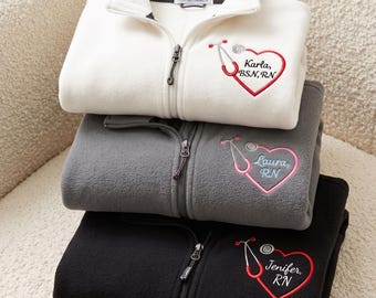 May include: A stack of three fleece zip-up jackets in white, grey, and black. Each jacket features an embroidered design on the chest consisting of a red or pink heart shaped like a stethoscope with the names Karla, Laura, or Jenifer and credentials.