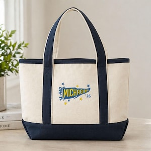 May include: A cream-colored canvas tote bag with navy blue trim and handles. The front features a blue pennant graphic with the yellow text MICHAGIN and the number '26, surrounded by small yellow and blue stars printed on the fabric.