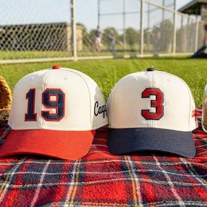 Könnte beinhalten: Zwei Baseballkappen mit cremefarbenen Kronen, roten und marineblauen Schirmen und marineblauen Zahlen. Eine Kappe zeigt die Zahl "19", die andere "3". Sie liegen auf einer rot-schwarz karierten Decke.