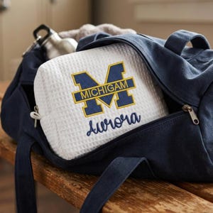 May include: A white waffle-textured zippered pouch featuring an embroidered navy blue and yellow block M logo with the word MICHIGAN inside, and the name Aurora embroidered in navy blue script below, tucked inside a navy blue canvas duffel bag.