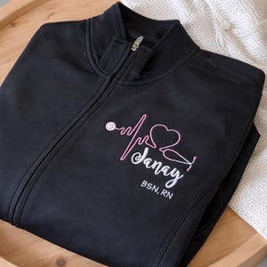 May include: Black zip-up jacket with a high collar. The jacket features a pink and white embroidered design of a heartbeat, stethoscope, and the name "Danay" with "BSN, RN" below. The jacket is folded on a wooden tray.