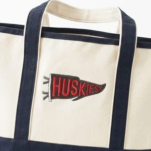 May include: Cream-colored canvas tote bag with navy blue accents and handles. The bag features a red and black pennant with the word "Huskies" embroidered on it. The bag is designed for carrying items.