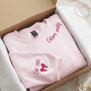 Puede incluir: Una sudadera de cuello redondo rosa claro con bordado rosa oscuro. El área del pecho muestra el texto Cutson sority, mientras que la manga presenta el texto Est. 1885 junto a un pequeño icono de lazo bordado.