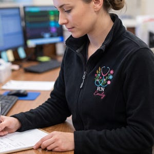 Könnte beinhalten: Eine schwarze Fleecejacke mit Reißverschluss und einer Stickerei auf der linken Brust. Das Motiv zeigt ein hellblaues Stethoskop, das mit bunten Blumen verziert ist, sowie die Buchstaben RN und den Namen Emily in rosa Schreibschrift.