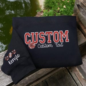 May include: Black sweatshirt with the word "CUSTOM" in red and white block letters. Below it, the words "Custom Text" are written in a cursive font. The left sleeve has the name "Kenzie" embroidered in white, along with a football and pom-pom design.