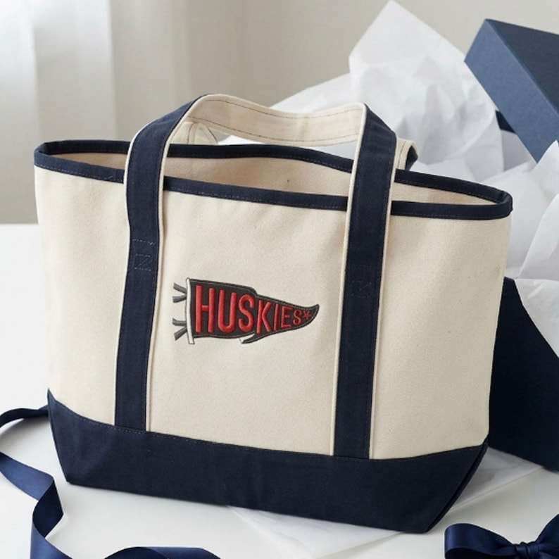 Natural/Navy Canvas tote bag with navy trim and handles, featuring embroidered college pennant design reading “HUSKIES” in bold red lettering, styled in a classic school spirit theme.