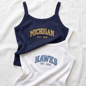 May include: A navy blue tank top with the gold text MICHIGAN EST. 1868 and a white tank top with the blue text HAWKS EST. 2026. Both garments feature thin shoulder straps, a scoop neckline, and collegiate-style lettering printed on the chest.