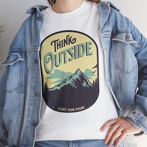 Op de afbeelding: Wit T-shirt met de tekst "Think Outside" en een berglandschap. Het shirt wordt gedragen met een lichtblauwe spijkerjas.