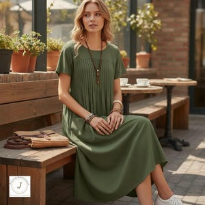 May include: An olive green midi dress with short sleeves and a pleated bodice. The dress has a round neckline and pockets. The model is wearing a long beaded necklace and bracelets. A straw bag sits on a wooden bench. The dress is a casual, everyday style.