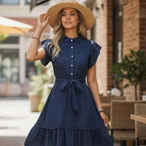 Puede incluir: Un vestido azul marino con lunares blancos, con cuello camisero, mangas cortas y cintura con cinturón. El vestido presenta un dobladillo con volantes y se combina con un sombrero de paja y zapatos de esparto. El vestido está abotonado por delante.