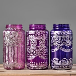 Puede incluir: Tres tarros de vidrio con diseños pintados en plata sobre fondo morado y azul. Los tarros están decorados con intrincados patrones.