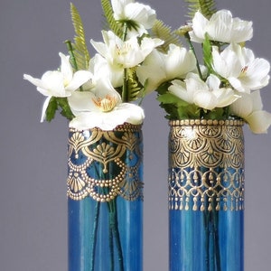 Puede incluir: Dos jarrones de vidrio azul con dise&ntilde;os pintados en oro, llenos de flores blancas y follaje verde.