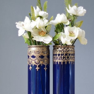 May include: Two tall, cylindrical blue vases with gold ornate designs, holding white flowers with yellow centers and green foliage. The vases are decorated with intricate gold patterns at the top and bottom.
