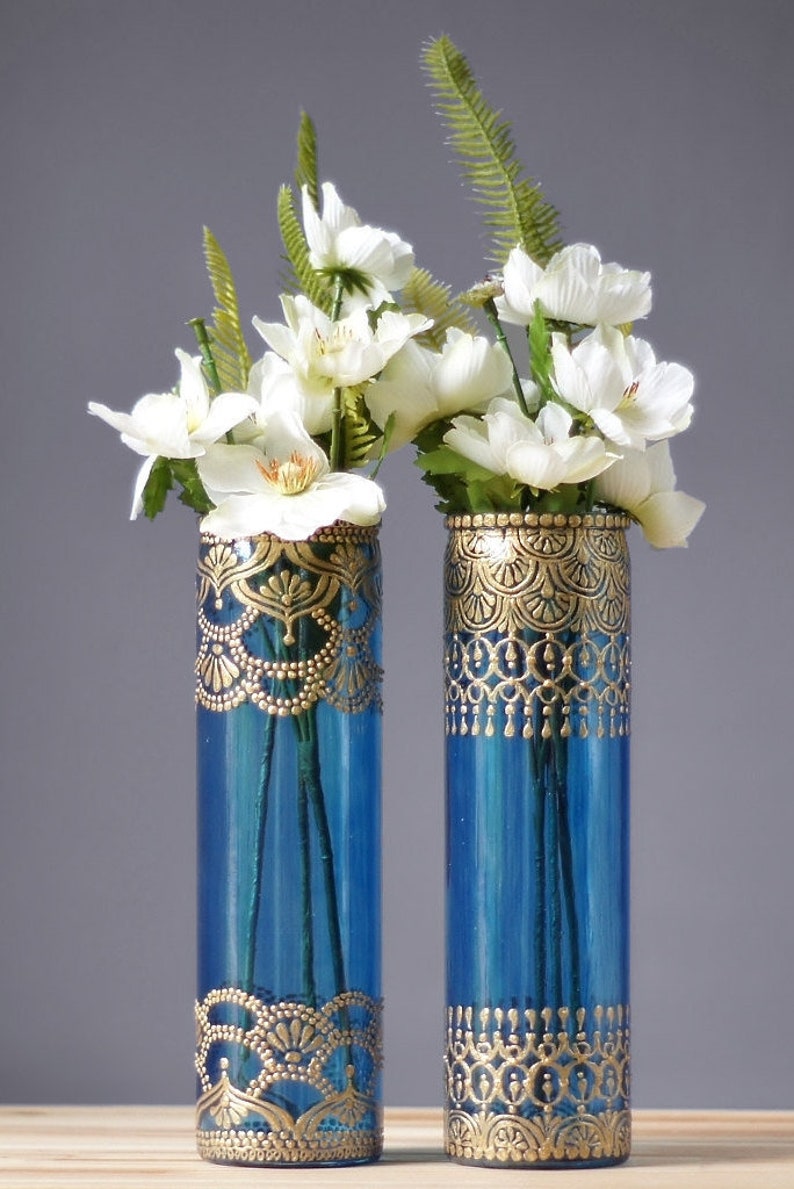 Puede incluir: Dos jarrones de vidrio azul con dise&ntilde;os pintados en oro, llenos de flores blancas y follaje verde.