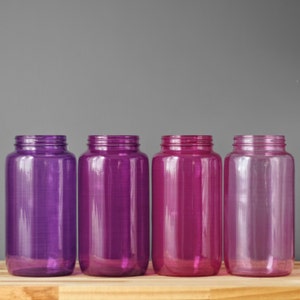 Può includere: Quattro bottiglie di vetro con un gradiente di colori viola e rosa. Le bottiglie sono allineate in una fila su una superficie di legno.