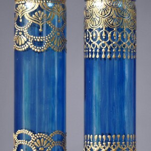 Op de afbeelding: Twee blauwe glazen vazen met gouden beschilderde ontwerpen. De vazen zijn cilindrisch en hebben een decoratief patroon rond de boven- en onderkant.