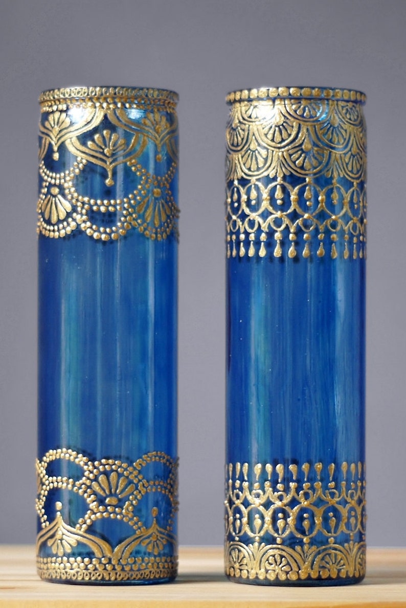 Op de afbeelding: Twee blauwe glazen vazen met gouden beschilderde ontwerpen. De vazen zijn cilindrisch en hebben een decoratief patroon rond de boven- en onderkant.
