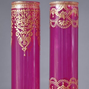 May include: Two magenta glass vases with gold painted designs. The vases are cylindrical and have a decorative gold band around the top and bottom.