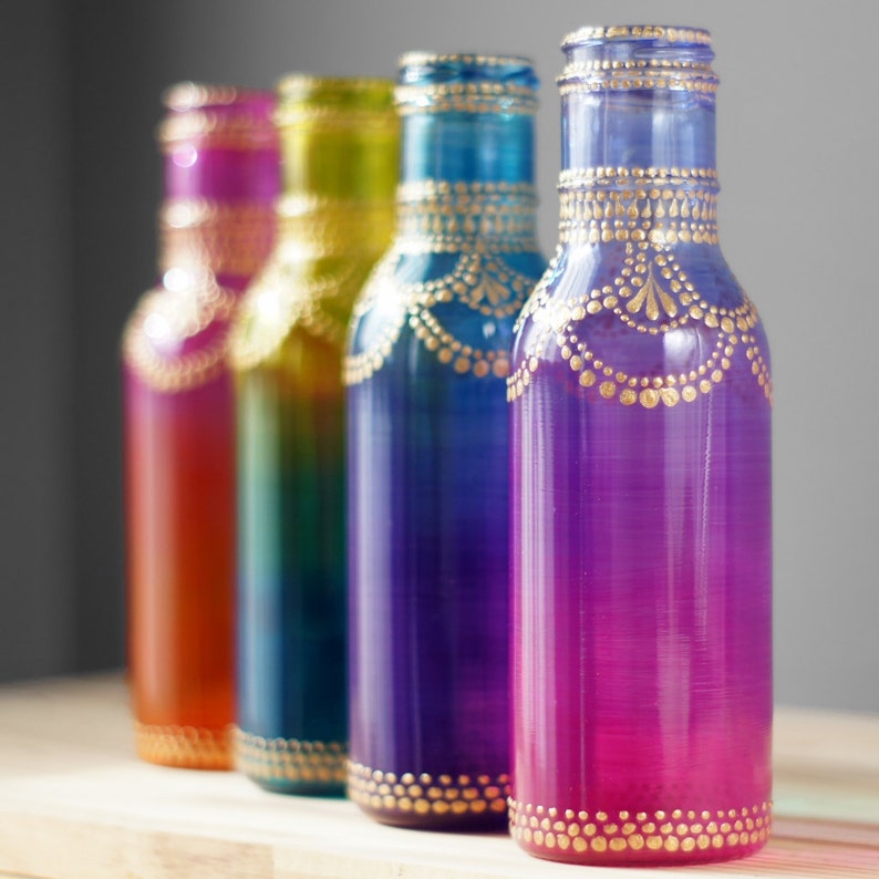 Op de afbeelding: Vier glazen flessen met gouden geschilderde ontwerpen. De flessen zijn geschilderd in tinten oranje, blauw, paars en roze.