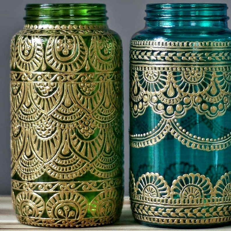 May include: Two glass jars with gold painted designs. The jar on the left is green glass with a gold floral pattern. The jar on the right is teal blue glass with a gold lace pattern.