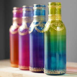 Op de afbeelding: Vier glazen flessen met ombre-verf en gouden accenten. De flessen zijn in een rij van links naar rechts gerangschikt, waarbij de kleuren overgaan van oranje naar roze, groen en blauw.