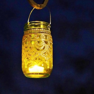May include: A decorative yellow glass jar lantern with intricate embossed patterns. The jar has a metal handle and a small lit candle inside, casting a warm glow. The lantern is suspended against a dark blue background.