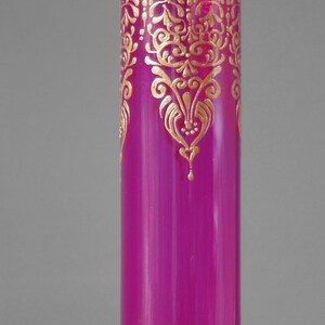 May include: A magenta glass vase with gold painted designs. The vase has a decorative pattern around the top and bottom.