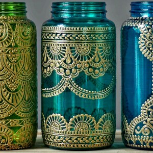 May include: Three glass jars with gold painted designs. The jars are teal, green, and blue. The designs are intricate and detailed, resembling henna or paisley patterns.