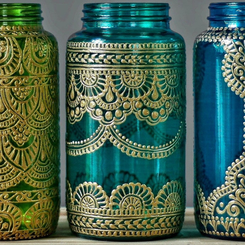 May include: Three glass jars with gold painted designs. The jars are teal, green, and blue. The designs are intricate and detailed, resembling henna or paisley patterns.