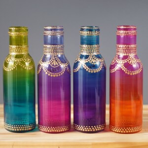 Op de afbeelding: Vier glazen flessen met ombre-verf in tinten van groen, paars, blauw en oranje. Elke fles heeft een gouden ontwerp geschilderd rond de boven- en onderkant.
