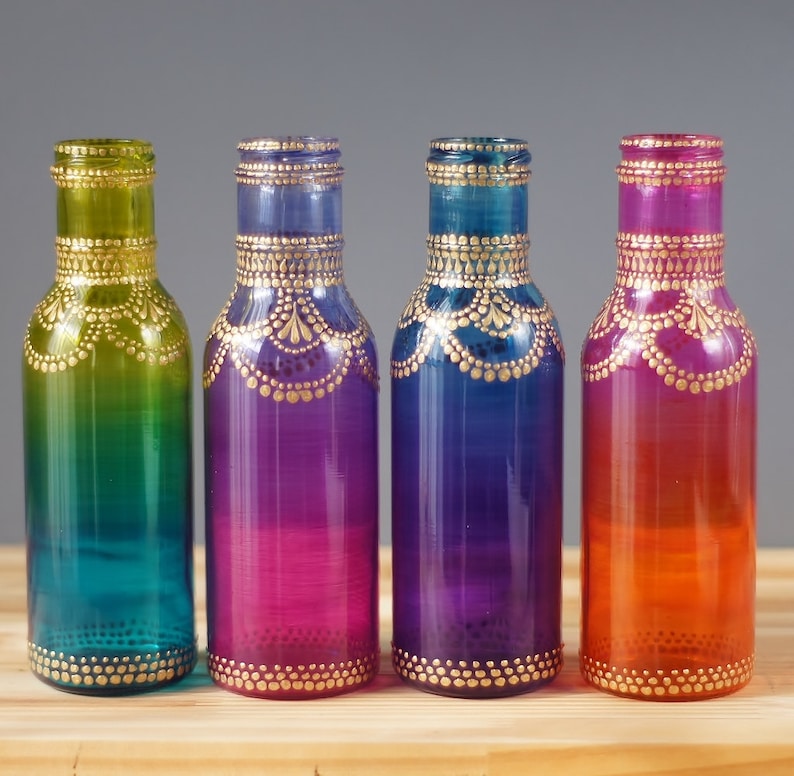 Op de afbeelding: Vier glazen flessen met ombre-verf in tinten van groen, paars, blauw en oranje. Elke fles heeft een gouden ontwerp geschilderd rond de boven- en onderkant.