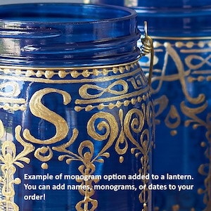 Op de afbeelding: Twee blauwe glazen potten met gouden beschilderde ontwerpen en een gouden draadhandvat. De pot aan de linkerkant heeft de letter "S" erop geschilderd. De tekst "Voorbeeld van een monogramoptie toegevoegd aan een lantaarn. Je kunt namen, monogrammen of datums aan je bestelling toevoegen!" is zichtbaar onderaan de afbeelding.