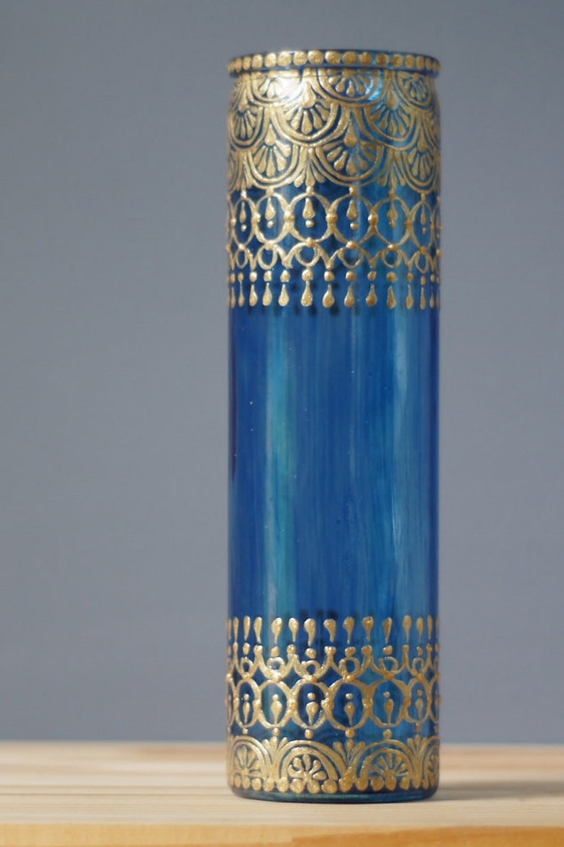 Op de afbeelding: Een blauwe glazen vaas met gouden beschilderde ontwerpen. De vaas heeft een cilindrische vorm en is versierd met ingewikkelde patronen.