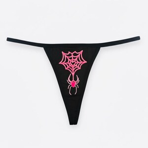 May include: Black thong underwear with a pink spider web and spider graphic. The spider web is centered on the front of the underwear, with the spider hanging below. The thong has thin black straps.