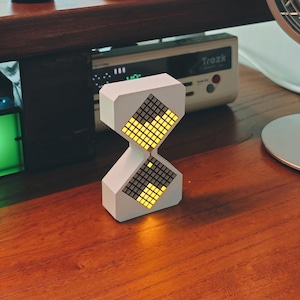 Puede incluir: Un reloj digital blanco con forma de reloj de arena que presenta dos pantallas LED en forma de diamante. Las secciones superior e inferior contienen una cuadrícula de píxeles cuadrados, algunos iluminados en amarillo y otros oscuros, creando un patrón geométrico.