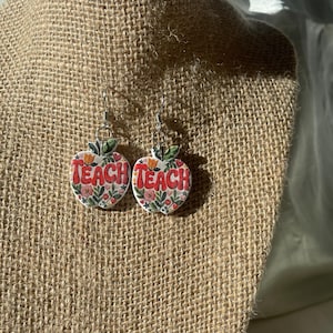 May include: A pair of apple-shaped earrings with the word "TEACH" in red letters. The earrings are decorated with floral patterns and green leaves. The earrings are hanging on a burlap display.