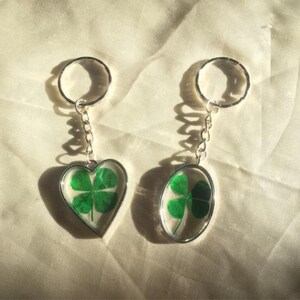 May include: Two keyrings with silver metal frames. One is heart-shaped, the other oval. Each contains a four-leaf clover preserved in clear resin. The keyrings are suspended from a chain and a silver ring.