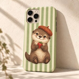 May include: A phone case featuring a light green and white vertical striped background with a brown otter wearing a rust-coloured beret and holding a small red strawberry. The case has a cutout for a triple-lens camera system on the top left.