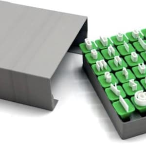 May include: A set of individual white letter and symbol stamps with green square bases, organised in a grid within a grey storage tray. The tray includes a matching grey sliding cover positioned to the left of the stamp collection.